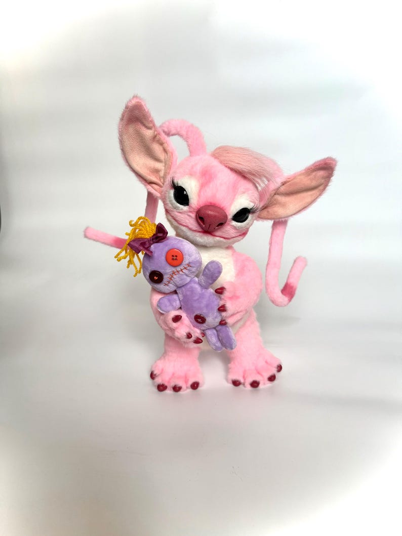 May include: A plush toy character with light pink fur, large ears, and long eyelashes, holding a smaller purple doll. The purple doll features yellow yarn hair, a dark purple bow, a single orange button eye, and stitched mouth details.