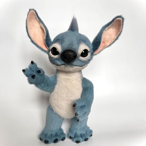 Puede incluir: Un personaje de peluche azul con orejas grandes, una nariz negra y ojos grandes. El personaje tiene un cuerpo azul claro, una barriga blanca y est&aacute; saludando con una mano. El peluche est&aacute; sobre una superficie blanca.