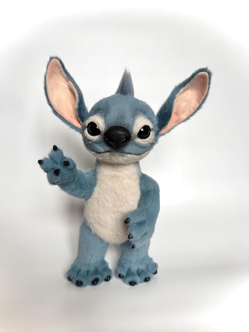 Puede incluir: Un personaje de peluche azul con orejas grandes, una nariz negra y ojos grandes. El personaje tiene un cuerpo azul claro, una barriga blanca y est&aacute; saludando con una mano. El peluche est&aacute; sobre una superficie blanca.