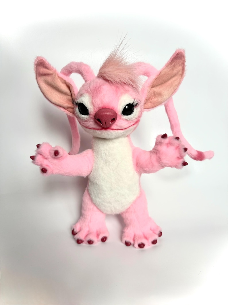 May include: A plush toy character with pink fur, a white belly patch, and large pointed ears. It features long eyelashes, a dark pink nose, and small dark claws on its hands and feet. A tuft of pink hair sits on its head, and it has two long, thin, trailing appendages extending from behind its ears.