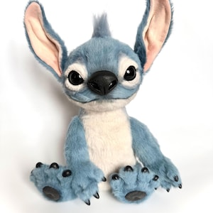 May include: A plush toy character with blue fur, large upright ears with light pink inner lining, and a cream-colored chest patch. The figure features large black eyes, a black nose, and black claws on its hands and feet, sitting in an upright position.