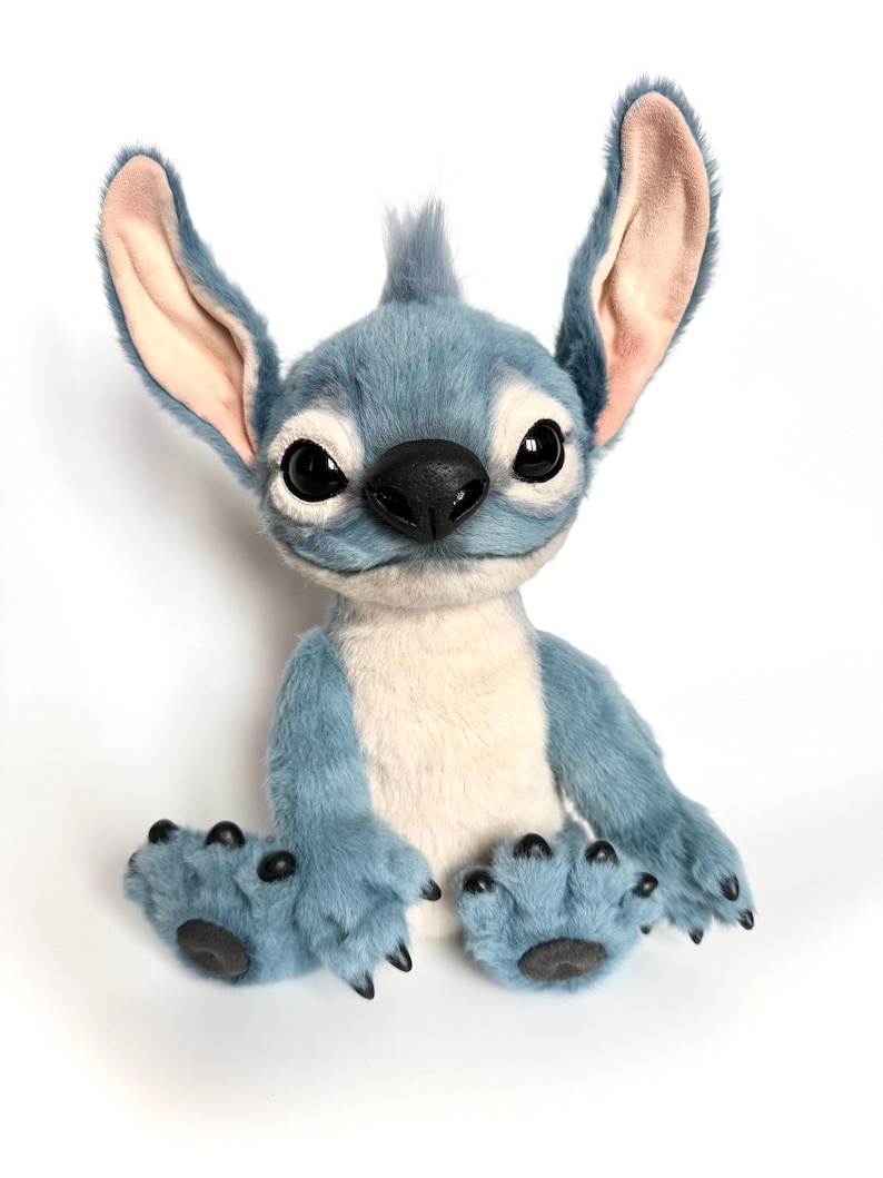 May include: A plush toy character with blue fur, large upright ears with light pink inner lining, and a cream-colored chest patch. The figure features large black eyes, a black nose, and black claws on its hands and feet, sitting in an upright position.