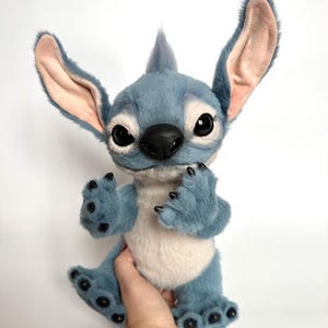 Puede incluir: Un personaje de peluche azul con orejas grandes, una nariz negra y ojos grandes. El peluche tiene un cuerpo azul claro, una barriga blanca y garras negras. El personaje es una figura animada popular.