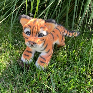 Op de afbeelding: Een klein, oranje en zwart tijgerwelpje knuffeldier met blauwe ogen, zittend in groen gras.