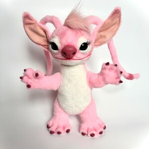 Puede incluir: Un peluche rosa con un personaje de grandes orejas, largas pesta&ntilde;as y un mech&oacute;n de pelo en la cabeza. El personaje tiene una barriga blanca, patas rosas y una boca peque&ntilde;a y sonriente. El juguete est&aacute; de pie con los brazos abiertos.
