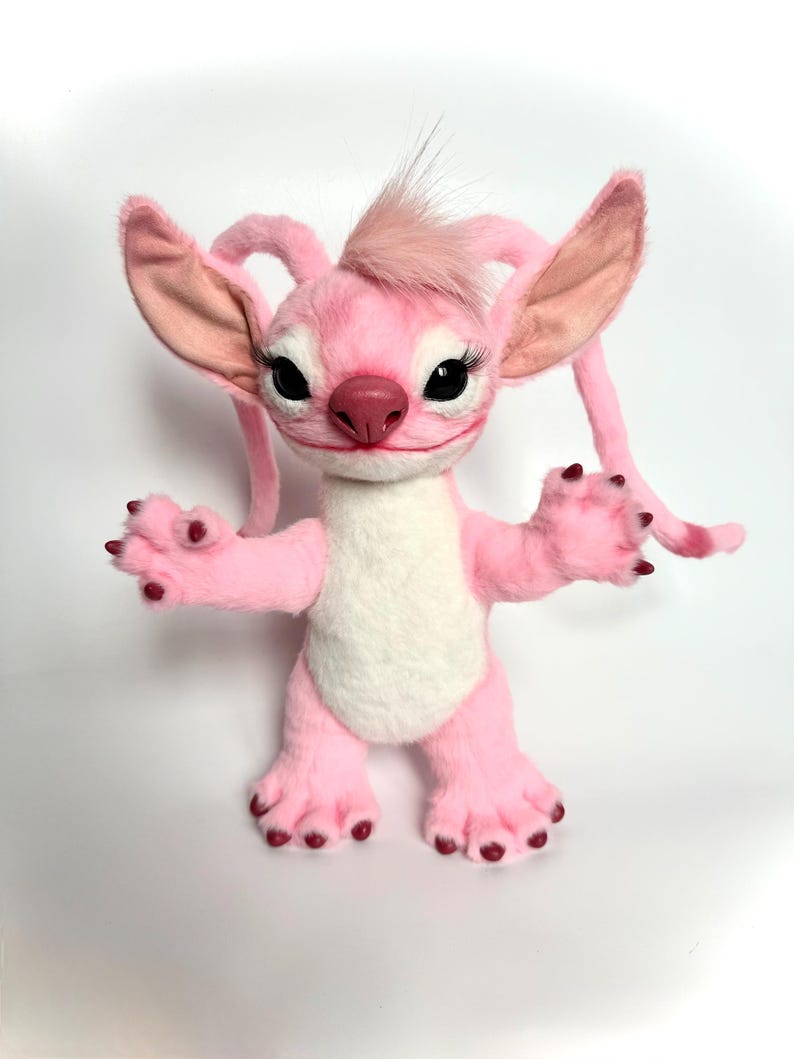Puede incluir: Un peluche rosa con un personaje de grandes orejas, largas pesta&ntilde;as y un mech&oacute;n de pelo en la cabeza. El personaje tiene una barriga blanca, patas rosas y una boca peque&ntilde;a y sonriente. El juguete est&aacute; de pie con los brazos abiertos.