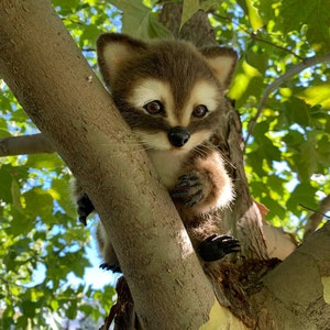 May include: A brown and white plush raccoon with black eyes and a black nose is sitting on a tree branch. The raccoon is looking to the side and has its paws resting on the branch.