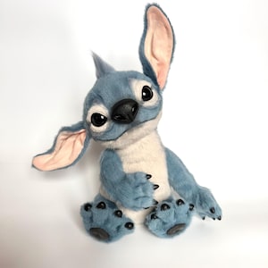 Puede incluir: Un peluche azul y blanco con grandes ojos expresivos y orejas desproporcionadas. El personaje tiene una nariz negra, garras negras y un mech&oacute;n de pelo en la cabeza. Est&aacute; sentado sobre una superficie blanca.