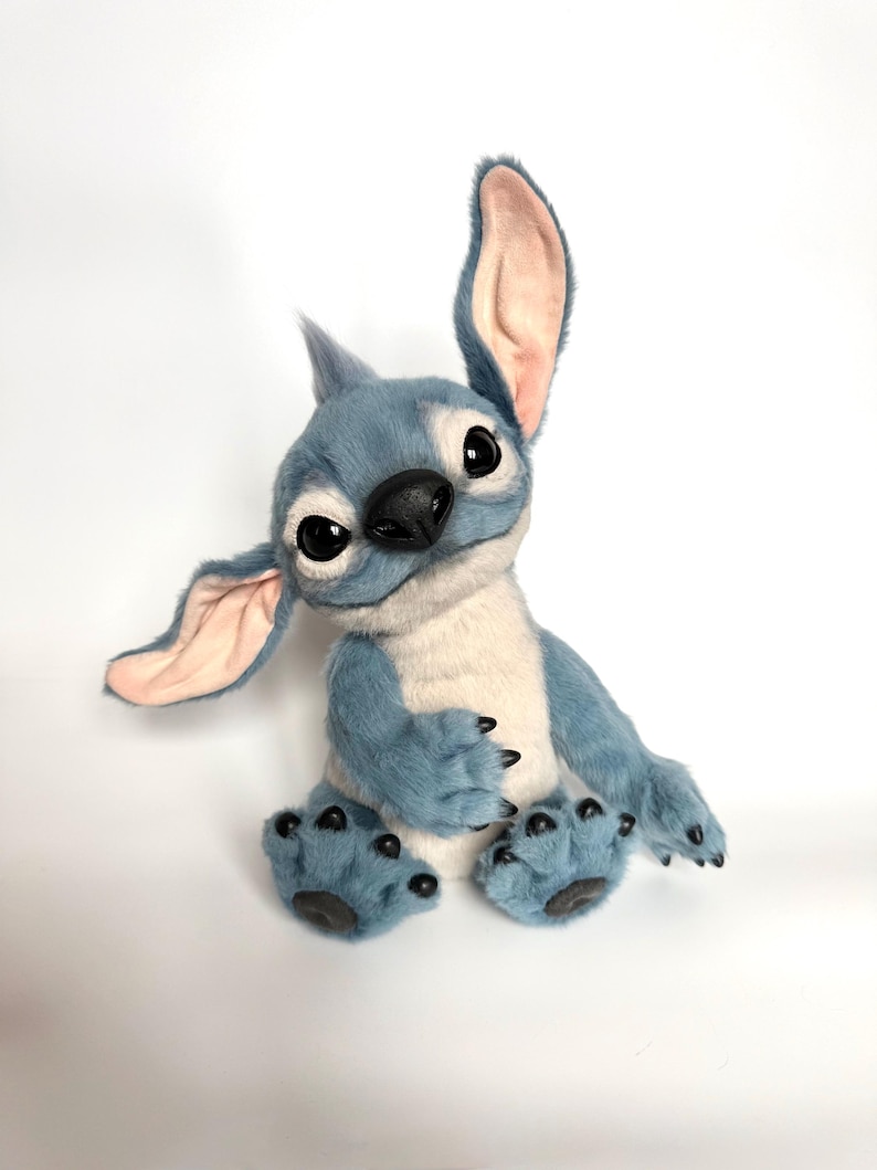 Puede incluir: Un peluche azul y blanco con grandes ojos expresivos y orejas desproporcionadas. El personaje tiene una nariz negra, garras negras y un mech&oacute;n de pelo en la cabeza. Est&aacute; sentado sobre una superficie blanca.
