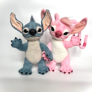 Puede incluir: Dos figuras de peluche de los personajes Stitch y Angel de la pel&iacute;cula animada Lilo & Stitch. Stitch es azul con orejas grandes, y Angel es rosa. Ambos tienen ojos grandes y est&aacute;n sobre una superficie blanca.