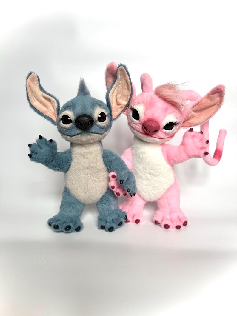 Puede incluir: Dos figuras de peluche de los personajes Stitch y Angel de la pel&iacute;cula animada Lilo & Stitch. Stitch es azul con orejas grandes, y Angel es rosa. Ambos tienen ojos grandes y est&aacute;n sobre una superficie blanca.
