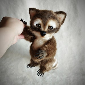 May include: A small, brown and white stuffed raccoon with black eyes and a black nose. The raccoon is sitting up with its paws raised, as if it is reaching out to someone.