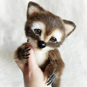 May include: A small, brown and white plush raccoon with black eyes and a black nose. The raccoon is being held in a person's hand.