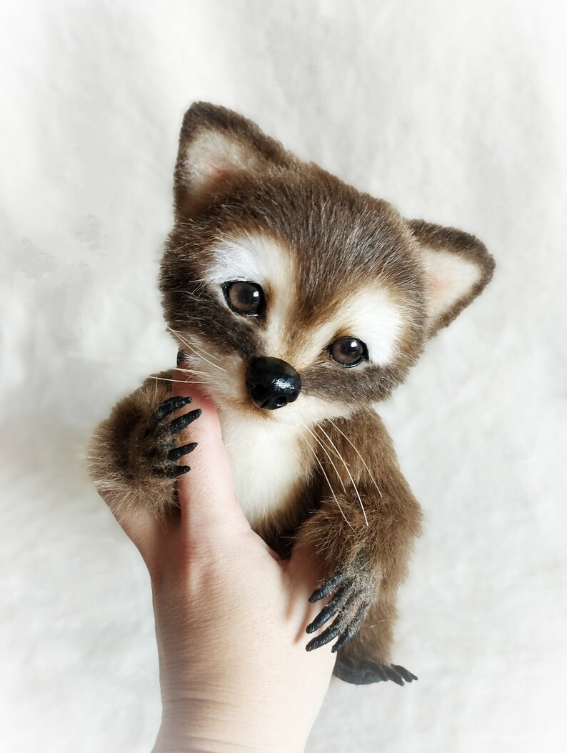 May include: A small, brown and white plush raccoon with black eyes and a black nose. The raccoon is being held in a person's hand.