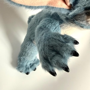 May include: A plush toy featuring blue fur with a soft texture, large ears with light pink inner lining, and dark, pointed claws on the feet. The creature has a rounded body and limbs covered in dense, synthetic blue fibers.
