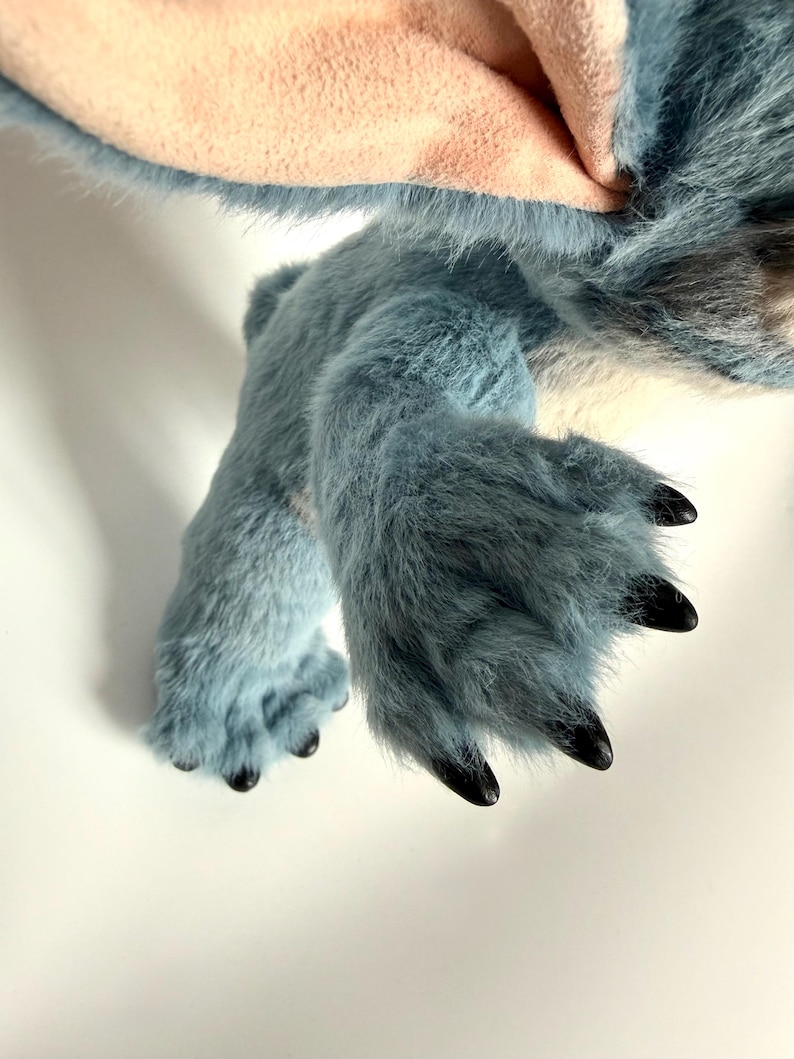 May include: A plush toy featuring blue fur with a soft texture, large ears with light pink inner lining, and dark, pointed claws on the feet. The creature has a rounded body and limbs covered in dense, synthetic blue fibers.
