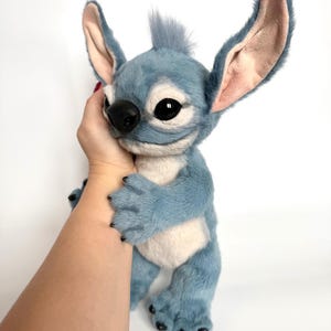 Puede incluir: Un peluche azul y blanco con orejas grandes, ojos negros y una nariz negra. El personaje tiene un mech&oacute;n de pelo en la cabeza y est&aacute; siendo sostenido por una mano. El juguete tiene una textura suave y peluda.