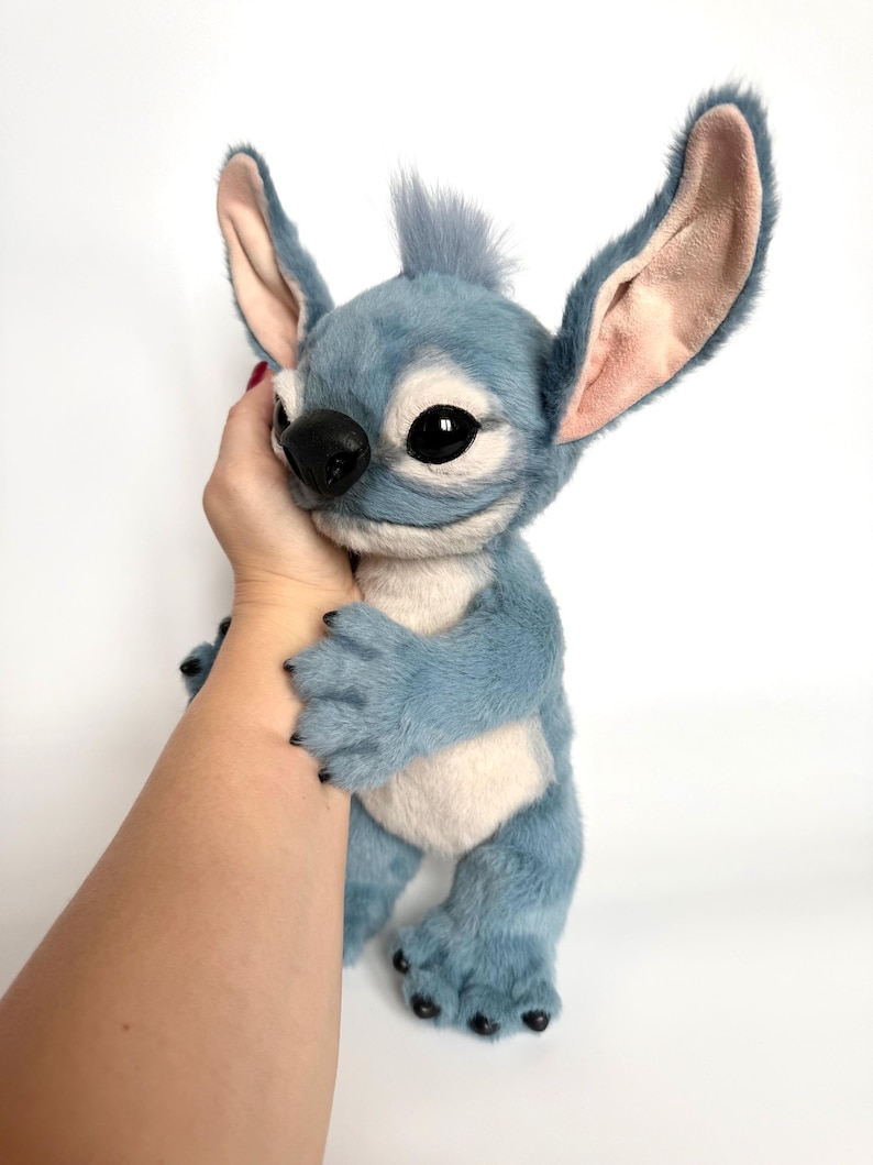 Puede incluir: Un peluche azul y blanco con orejas grandes, ojos negros y una nariz negra. El personaje tiene un mech&oacute;n de pelo en la cabeza y est&aacute; siendo sostenido por una mano. El juguete tiene una textura suave y peluda.