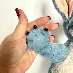 May include: A plush toy featuring blue fur, a light beige chest, and large ears with light pink inner lining. The toy has a blue paw with black claws extending from the arm. The texture of the fur is soft and dense, with visible stitching.