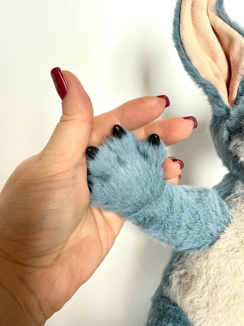 May include: A plush toy featuring blue fur, a light beige chest, and large ears with light pink inner lining. The toy has a blue paw with black claws extending from the arm. The texture of the fur is soft and dense, with visible stitching.