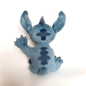 May include: A blue plush toy character with large pointed ears, a small tuft of hair on the head, and dark blue patterned markings on the back. The character is shown from behind, with one arm raised in a waving gesture and small black claws.