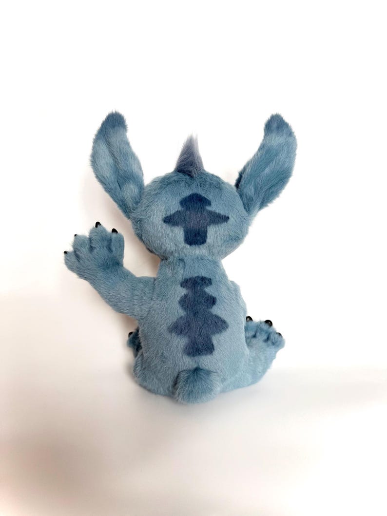 May include: A blue plush toy character with large pointed ears, a small tuft of hair on the head, and dark blue patterned markings on the back. The character is shown from behind, with one arm raised in a waving gesture and small black claws.