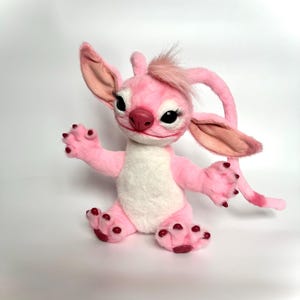 Puede incluir: Un peluche rosa con orejas grandes, un mech&oacute;n de pelo y una cola larga. El juguete tiene una barriga blanca, detalles en rojo oscuro en los pies y largas pesta&ntilde;as. El juguete est&aacute; sobre una superficie blanca.