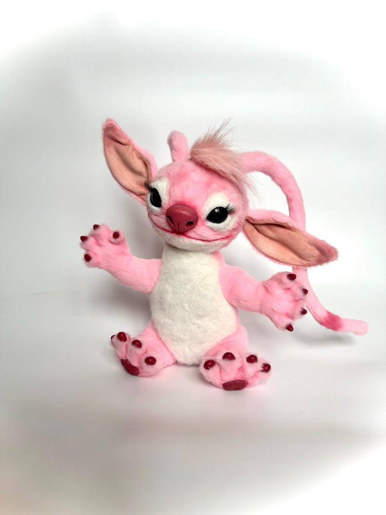 Puede incluir: Un peluche rosa con orejas grandes, un mech&oacute;n de pelo y una cola larga. El juguete tiene una barriga blanca, detalles en rojo oscuro en los pies y largas pesta&ntilde;as. El juguete est&aacute; sobre una superficie blanca.