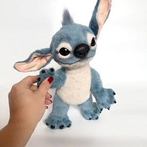 May include: A plush toy character with blue fur, large pointed ears with light pink inner lining, a cream-colored chest patch, and a black nose. The figure features large dark eyes, a small tuft of hair on top, and black claws on its hands and feet.