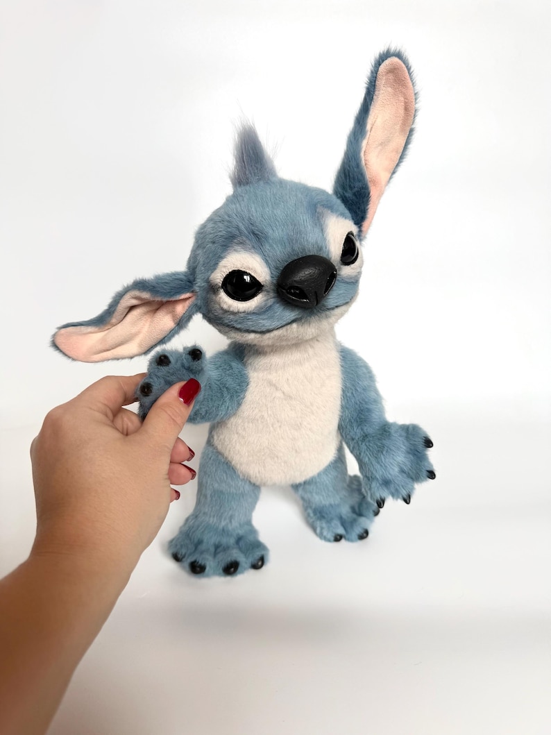 May include: A plush toy character with blue fur, large pointed ears with light pink inner lining, a cream-colored chest patch, and a black nose. The figure features large dark eyes, a small tuft of hair on top, and black claws on its hands and feet.