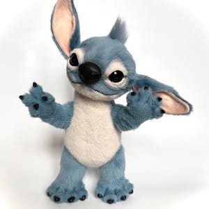 Puede incluir: Mu&ntilde;eco de peluche Stitch azul y blanco con orejas grandes, ojos negros y nariz negra. El mu&ntilde;eco tiene los brazos extendidos y est&aacute; de pie sobre sus pies. El pelaje es suave y detallado, con una expresi&oacute;n amigable.