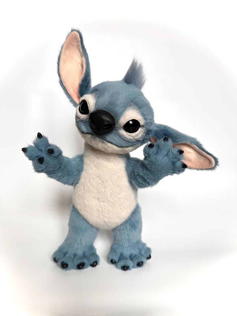 Puede incluir: Mu&ntilde;eco de peluche Stitch azul y blanco con orejas grandes, ojos negros y nariz negra. El mu&ntilde;eco tiene los brazos extendidos y est&aacute; de pie sobre sus pies. El pelaje es suave y detallado, con una expresi&oacute;n amigable.