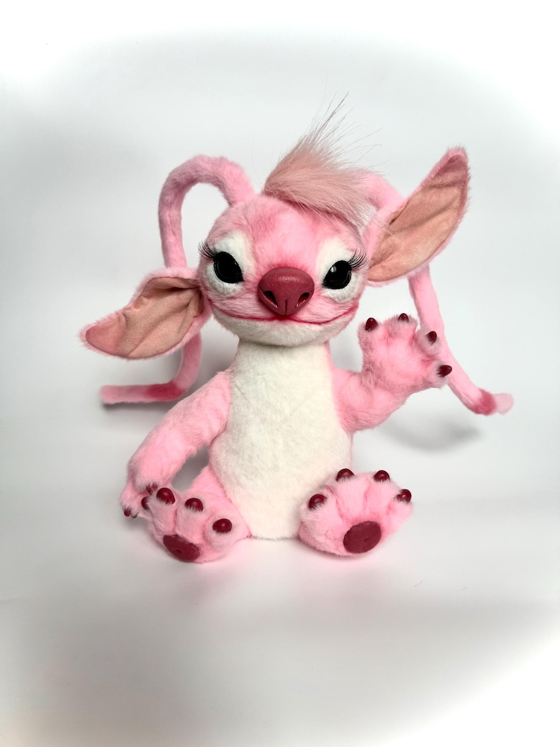 Puede incluir: Un personaje de peluche rosa con orejas grandes, largas pesta&ntilde;as y un mech&oacute;n de pelo en la cabeza. El personaje tiene una barriga blanca, patas rosas con almohadillas de color rojo oscuro y est&aacute; sentado con una pata levantada.