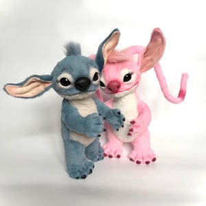 Puede incluir: Dos figuras de peluche de los personajes Stitch y Angel de la pel&iacute;cula animada Lilo & Stitch. Stitch es azul con orejas grandes, y Angel es rosa. Ambos tienen ojos negros y est&aacute;n en una pose de abrazo. Las figuras est&aacute;n sobre un fondo blanco.