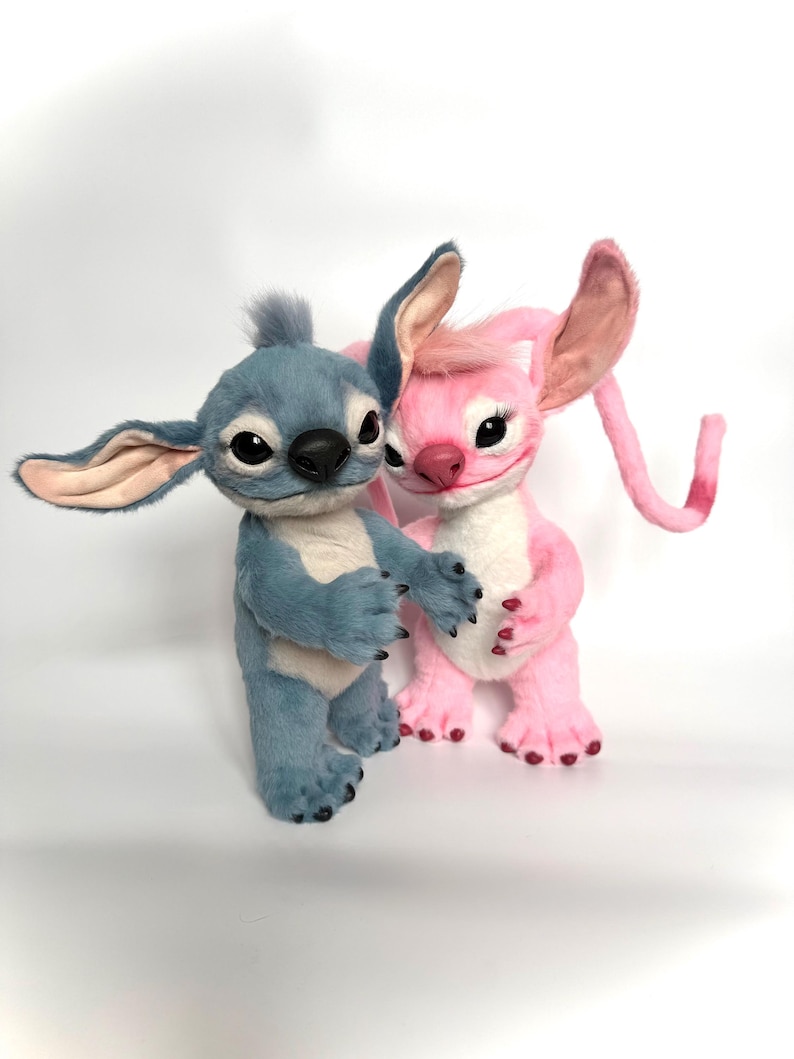 Puede incluir: Dos figuras de peluche de los personajes Stitch y Angel de la pel&iacute;cula animada Lilo & Stitch. Stitch es azul con orejas grandes, y Angel es rosa. Ambos tienen ojos negros y est&aacute;n en una pose de abrazo. Las figuras est&aacute;n sobre un fondo blanco.
