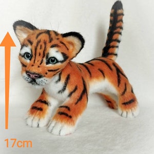Op de afbeelding: Een klein, oranje en zwart tijgerwelpje van pluche, staand op een wit oppervlak. Het speelgoed is 17 cm hoog.