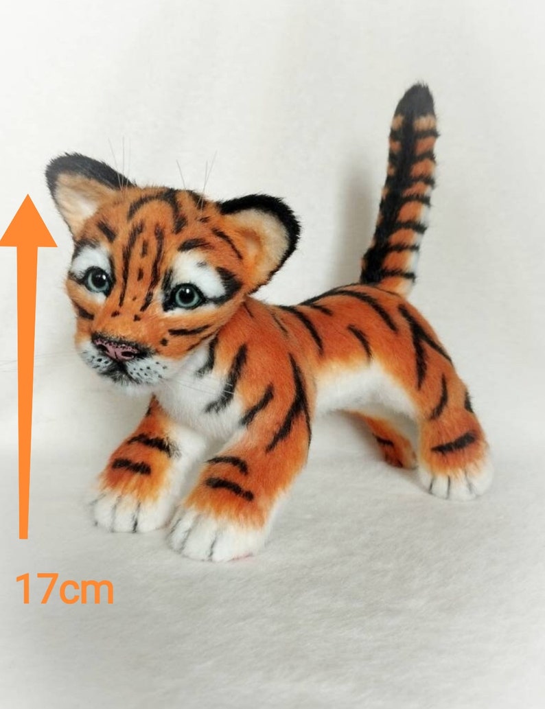 Op de afbeelding: Een klein, oranje en zwart tijgerwelpje van pluche, staand op een wit oppervlak. Het speelgoed is 17 cm hoog.