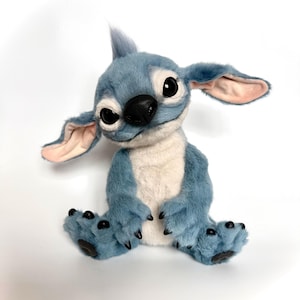 Puede incluir: Un peluche azul con orejas grandes, ojos negros y nariz negra. El peluche tiene vientre blanco y garras negras en los pies. La criatura est&aacute; sentada con las piernas extendidas y los brazos a los lados.