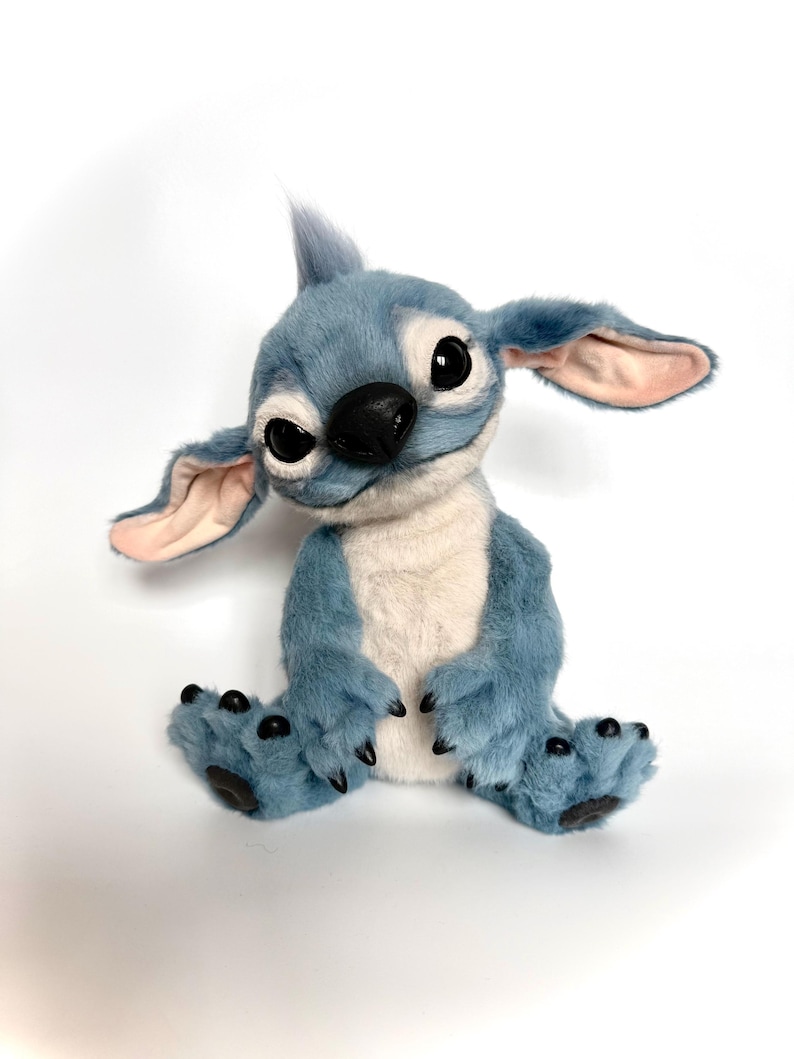 Puede incluir: Un peluche azul con orejas grandes, ojos negros y nariz negra. El peluche tiene vientre blanco y garras negras en los pies. La criatura est&aacute; sentada con las piernas extendidas y los brazos a los lados.