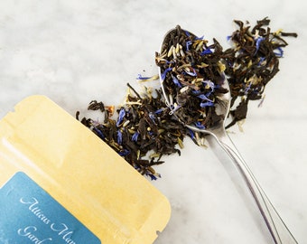 May include: A silver spoon filled with loose leaf tea, including black tea leaves, blue cornflower petals, and white pieces of dried fruit. The tea is scattered on a white marble surface next to a yellow pouch with the text "Atticus Manor Garden" printed on it.