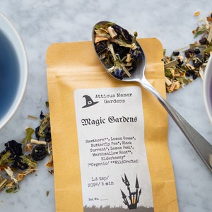 May include: A brown paper pouch with a label that reads "Atticus Manor Gardens" and "Magic Gardens". The label lists the ingredients of the tea blend, including Hawthorn, Lemon Grass, Butterfly Pea, Black Currant, Lemon Peel, Marshmallow Root, Elderberry, and notes that it is organic and wildcrafted. The pouch is filled with loose leaf tea, and a silver spoon is holding some of the tea leaves. There are two cups of blue and purple tea in the background.