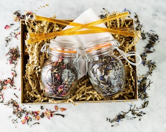 May include: Two glass jars filled with loose leaf tea, with orange lids and silver clasps. The jars are sitting in a brown box with shredded paper and two yellow wooden tea stirrers.
