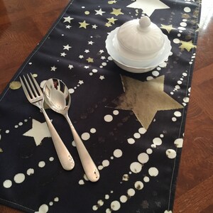 May include: A black table runner with a white and gold star pattern. The runner is decorated with white polka dots and has a subtle sheen. A white sugar bowl with a lid is placed on the runner.