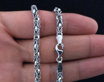 Op de afbeelding: Een zilveren ketting met een delicaat, ingewikkeld ontwerp. De ketting wordt op een hand getoond, en de sluiting is zichtbaar.