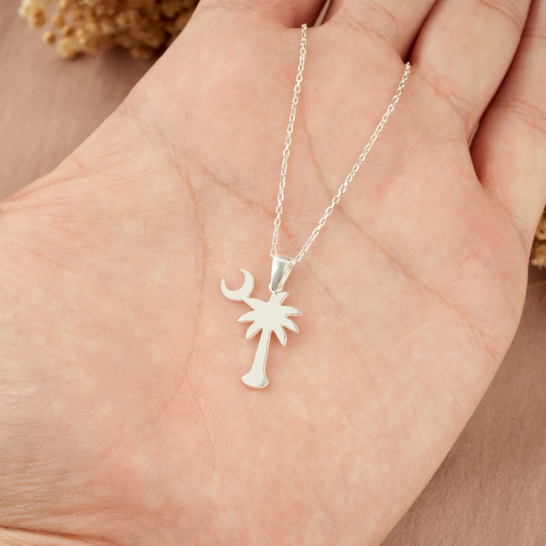Op de afbeelding: Een zilveren ketting met een palmboom en halve maan hanger. De ketting ligt op een hand.