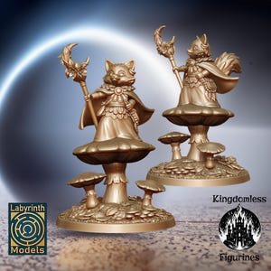 May include: Two golden painted figurines of fox-like creatures standing on mushroom bases. The figures are holding staffs with crescent moon tops. The bases are decorated with leaves and mushrooms. The figures are labeled "Kingdomless Figurines" and "Labyrinth Models".