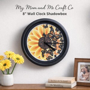 Puede incluir: Un reloj de pared de unos 20 cm de diámetro con marco negro. La esfera presenta un diseño de girasol en capas en amarillo y naranja, con recortes de mariposas negras. El texto "My Mom and Me Craft Co" está en la parte superior.