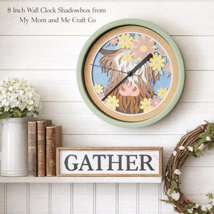 Puede incluir: Un reloj de pared de aproximadamente 20 cm de diámetro, con un marco verde y un diseño en capas que presenta una vaca de las Tierras Altas y flores. Las manecillas del reloj son negras. La palabra "GATHER" está en un letrero de madera debajo.