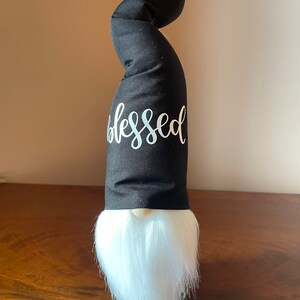Puede incluir: Un gnomo negro con barba blanca y un sombrero negro con la palabra "blessed" escrita en blanco en la parte delantera.