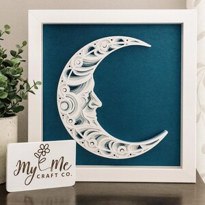 May include: White framed artwork featuring a layered paper crescent moon with a face design against a teal background. The artwork is displayed on a dark surface with a small white sign that reads "my & me CRAFT CO."